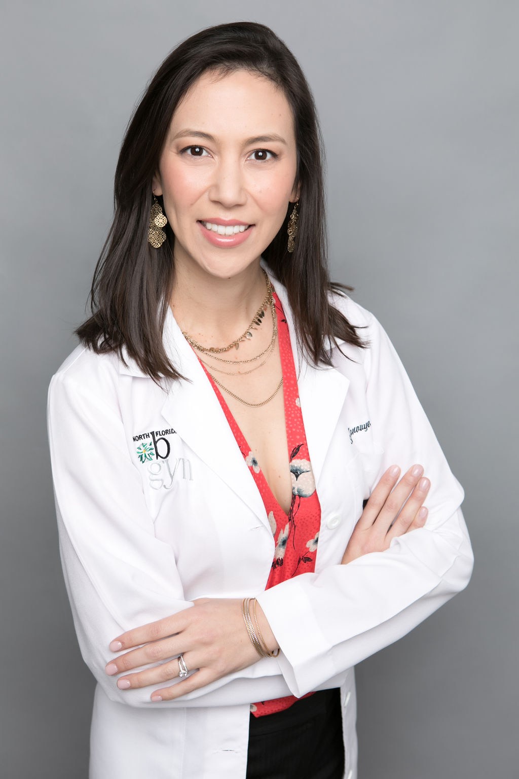 Dr. Staci Tanouye standing with crossed arms in a white lab coat, red floral blouse, black bottoms, and gold jewelry.