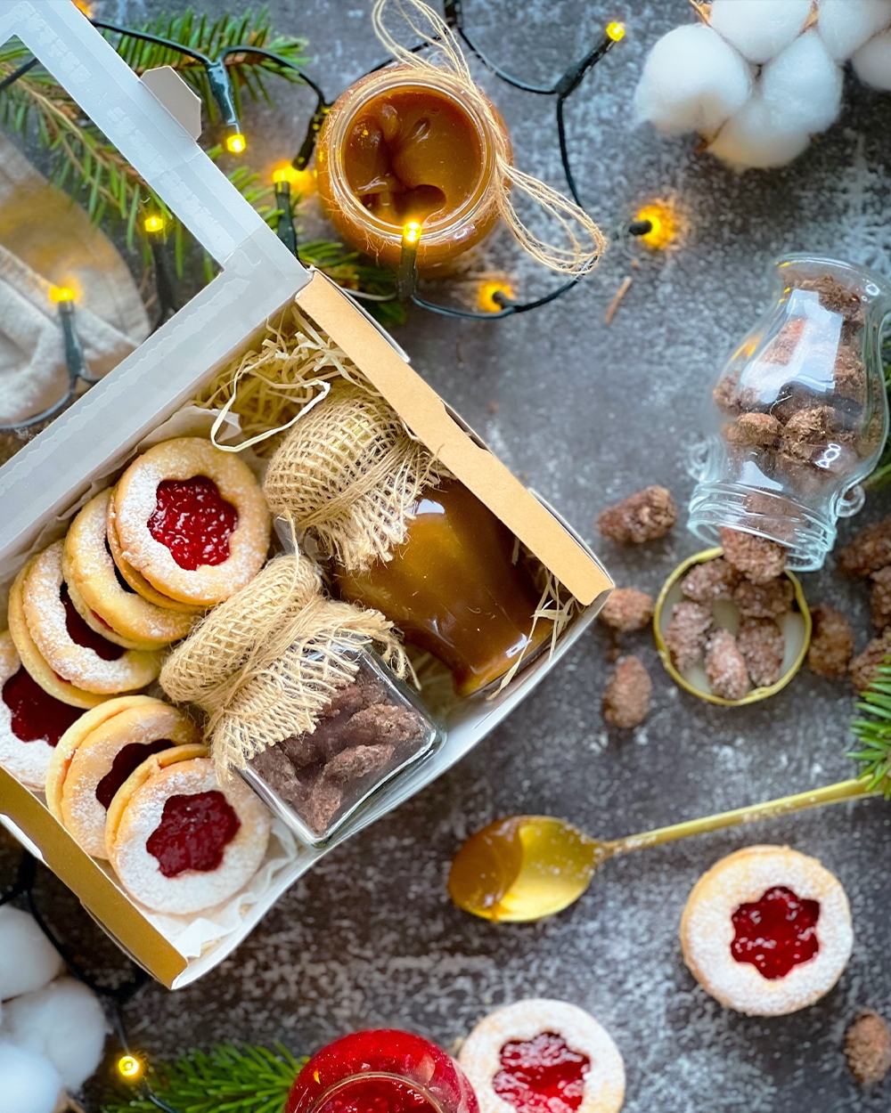 Box of fruit-filled sugar cookies with powdered sugar sprinkled on top next to jarred sauces and a gold spoon. 