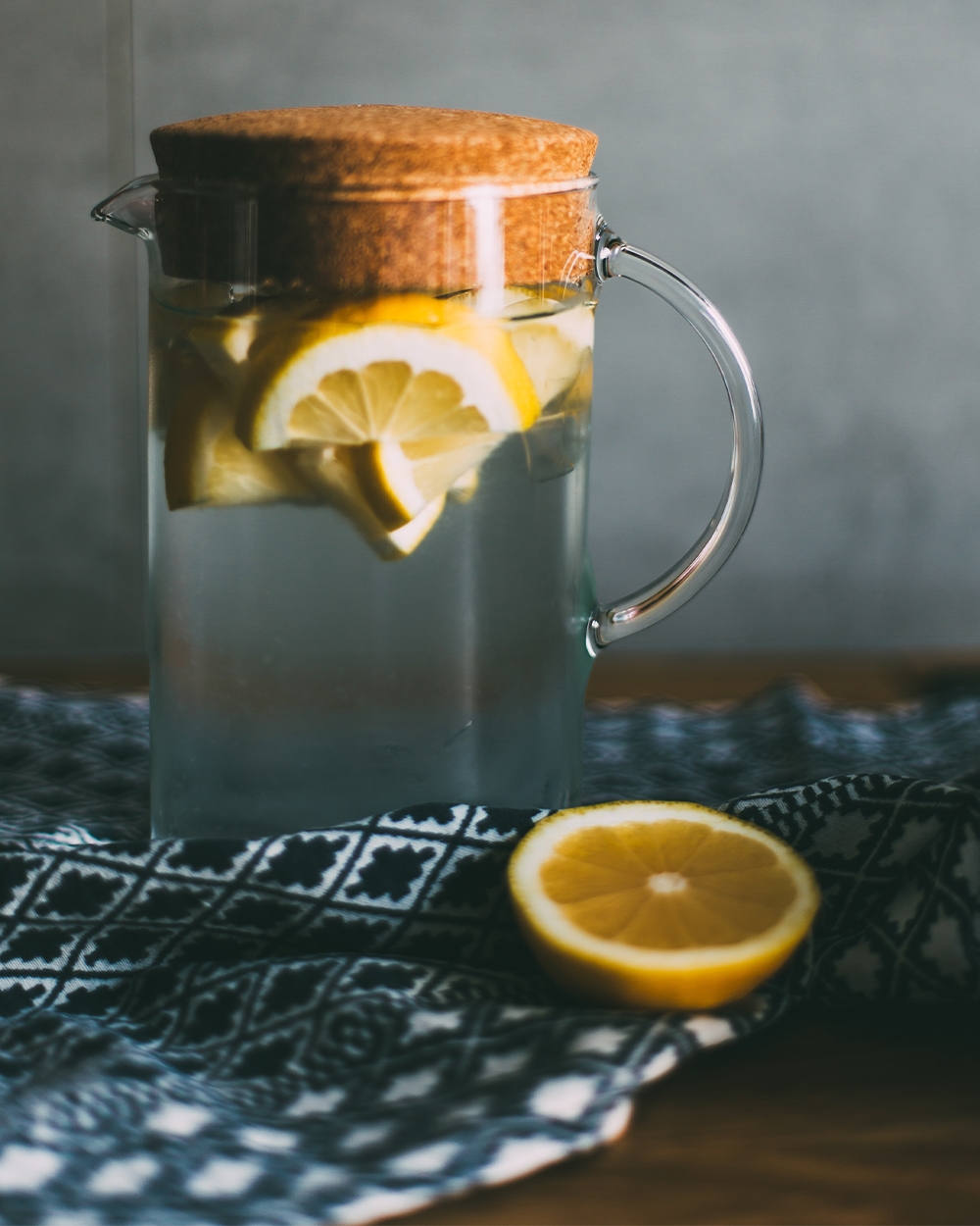 Glass pitcher of lemon water with sliced lemons and a sealed, cork top sitting on a black and white table runner.