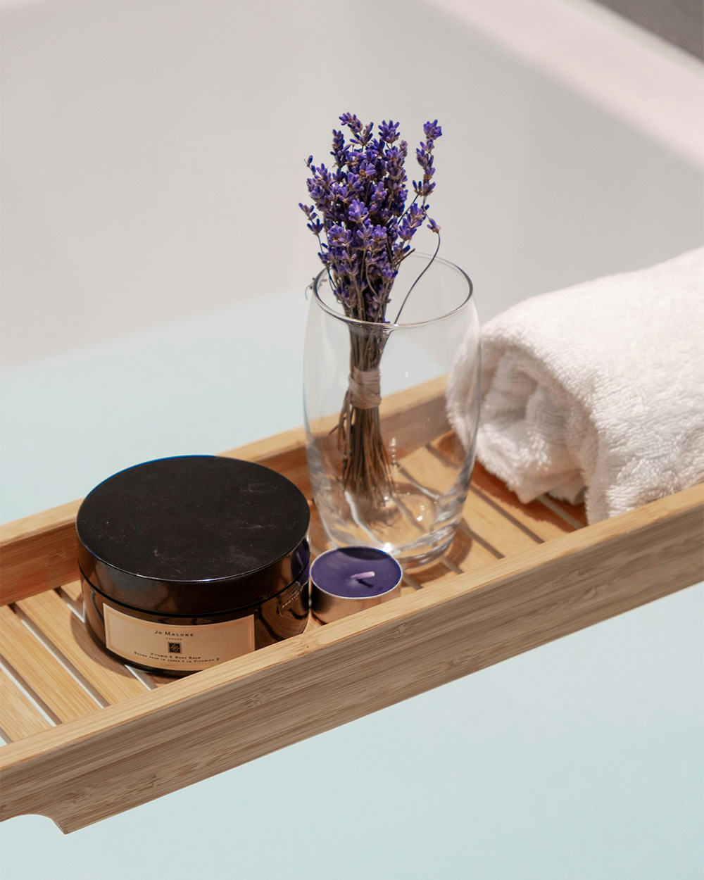 Wooden bath rack with jarred skincare, purple candle, glass vase with lavender sprigs, and rolled white towel.