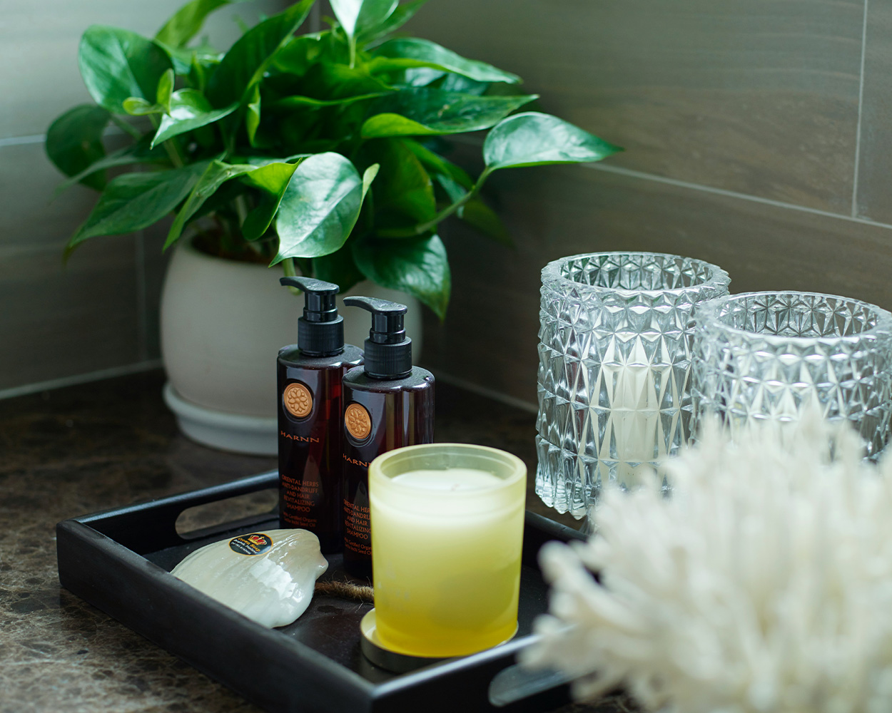 Bathroom counter with black tray holding a candle, soaps and lotions, and two crystal glasses sitting next to a potted plant.