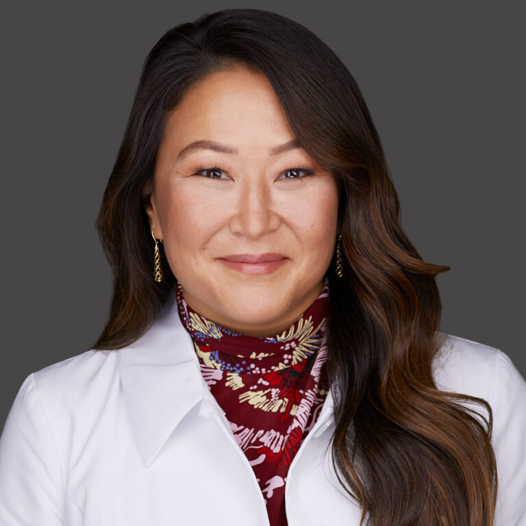 A smiling brunette woman wearing a floral turtleneck top and white lab coat.