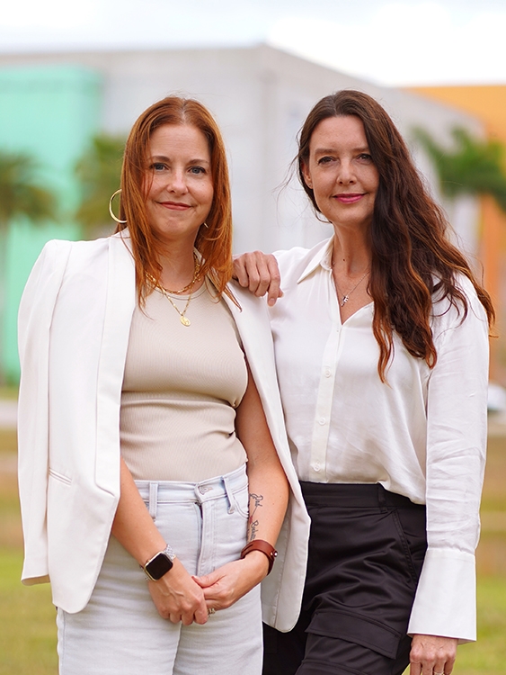 Red-headed woman wearing light tan tank top, white blazer, and lightwash jeans posing with dark-haired woman wearing white blouse and black slacks.
