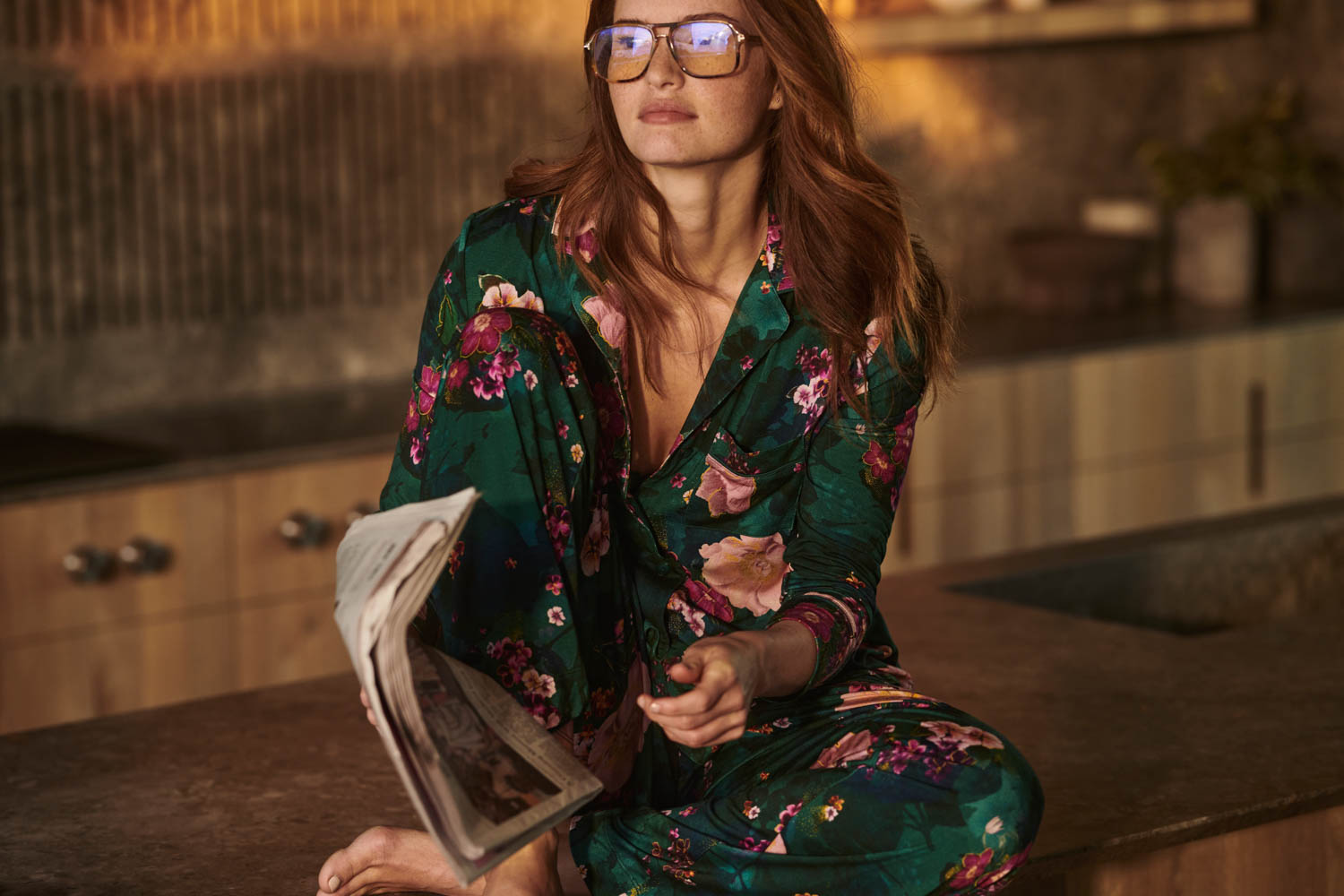 Soma<sup class=st-superscript>®</sup> model sitting on a kitchen counter, wearing dark green floral women’s pajamas and large aviator eyeglasses.