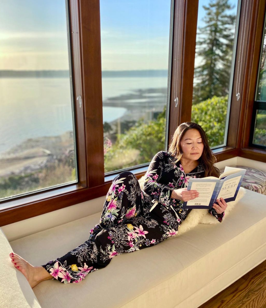 A brunette woman wearing floral pajamas while reading a book and lounging on a couch.