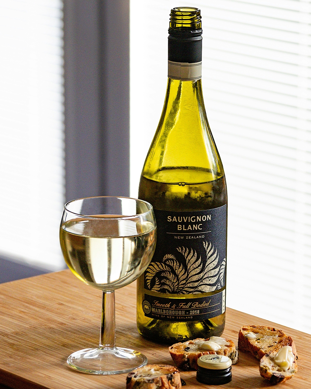 Open bottle of Sauvignon Blanc, full glass of wine, small buttered bread bites on a wooden table.