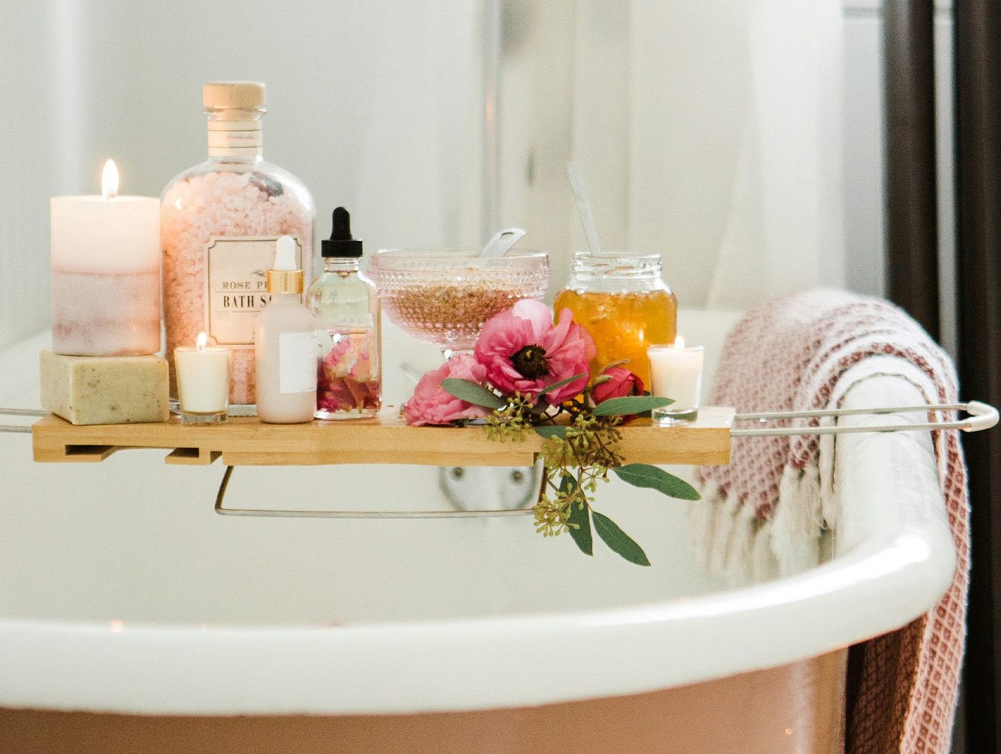 Lit candles, bath salts, soap, and flowers sitting on a wooden bath rack on a bathtub.