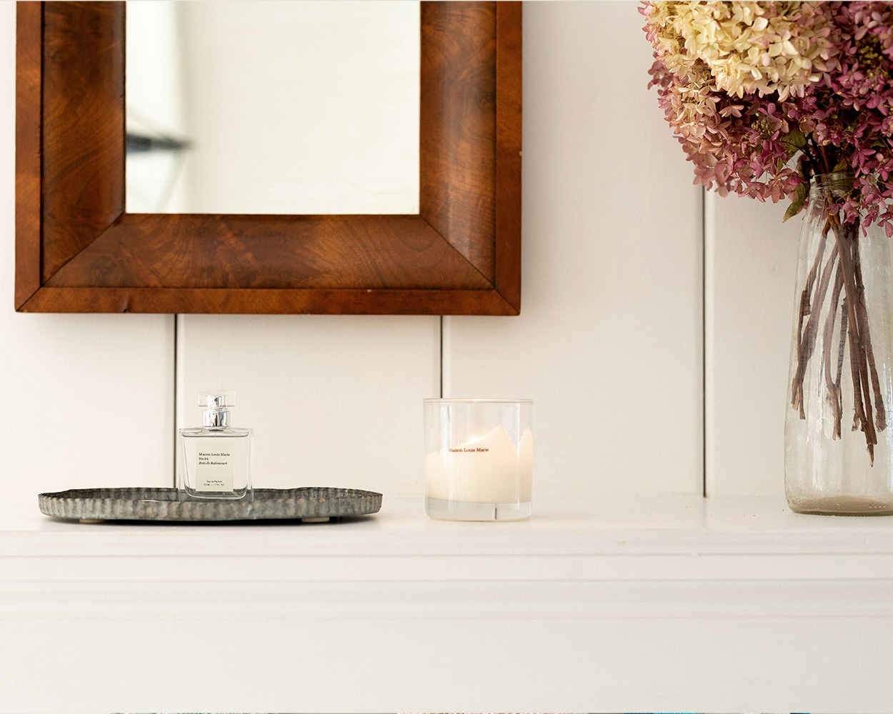 Wooden framed mirror hung on a wall above a shelf with a metal tray, perfume bottle, candle, and vase with flowers inside.