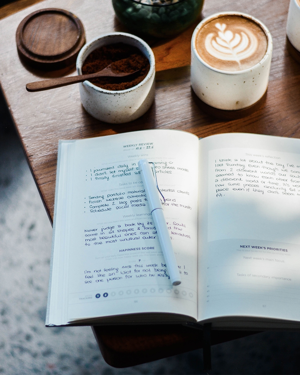 Open journal with handwritten notes and closed pen on top of a wooden table, next to a mug of coffee.