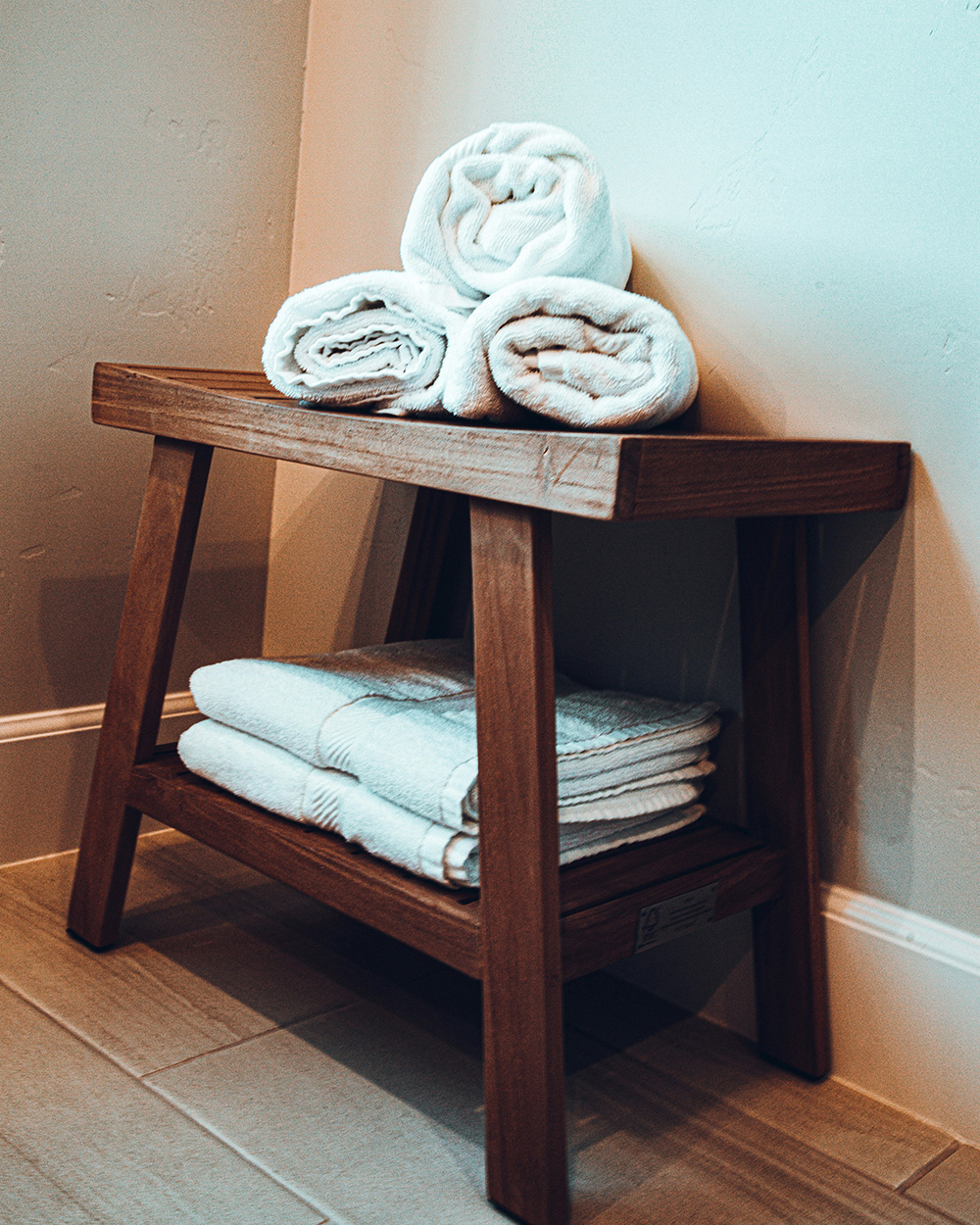Wooden bath stool with storage for towels, white towels folded underneath, and white towels rolled and organized on top.