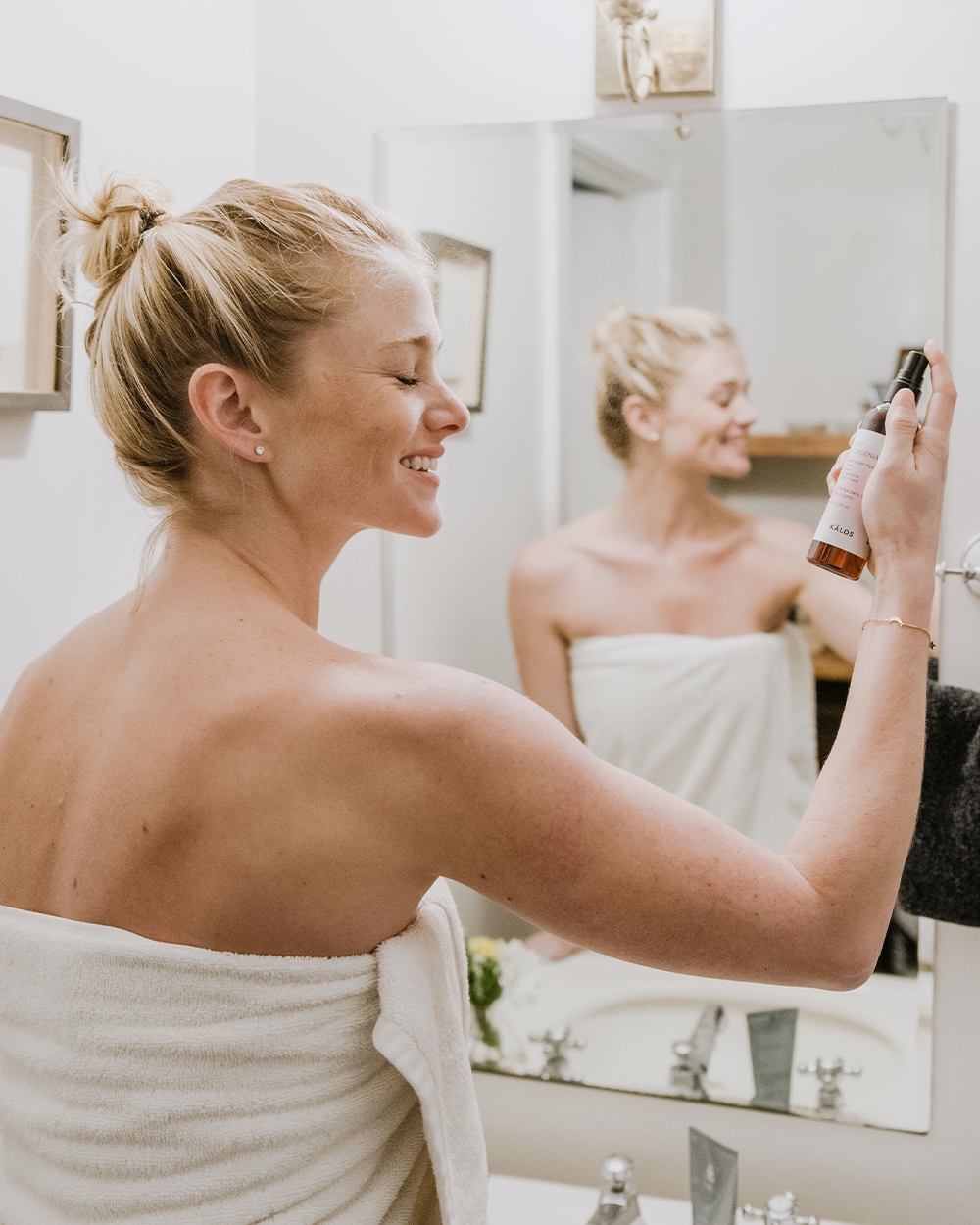  Model standing in front of bathroom sink wearing a white towel and spraying skincare on her face.