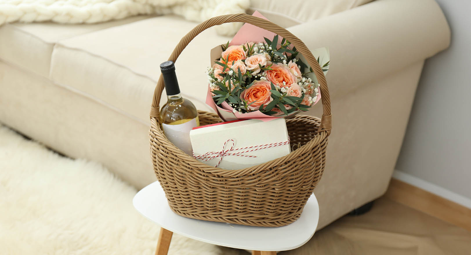 A photo of a wicker gift basket with flowers sitting on a small white table in front of a white couch in a living room.