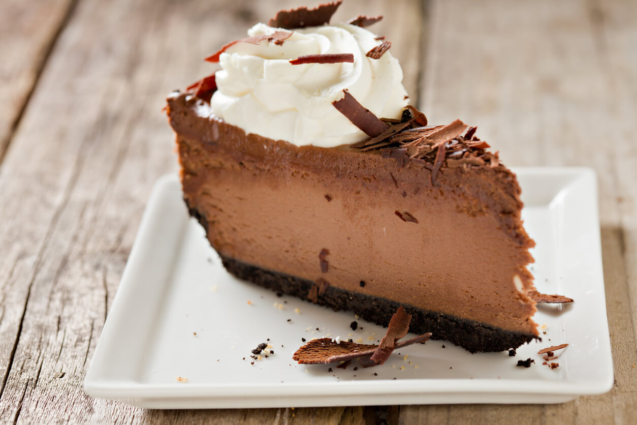 A high angle close up of a square white plate with a fresh slice of chocolate cheesecake with a dab of whipped cream and topped with chocolate shavings. Shot on a grungy old wooden picnic table.