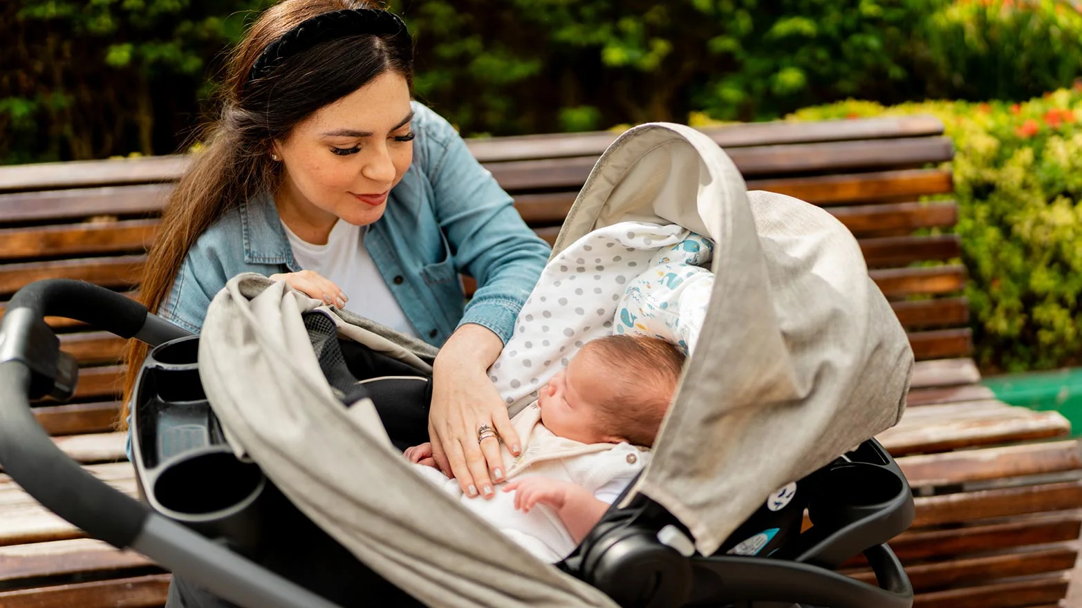 A stock photo of a woman sitting on a bench and looking at a baby in a stroller.