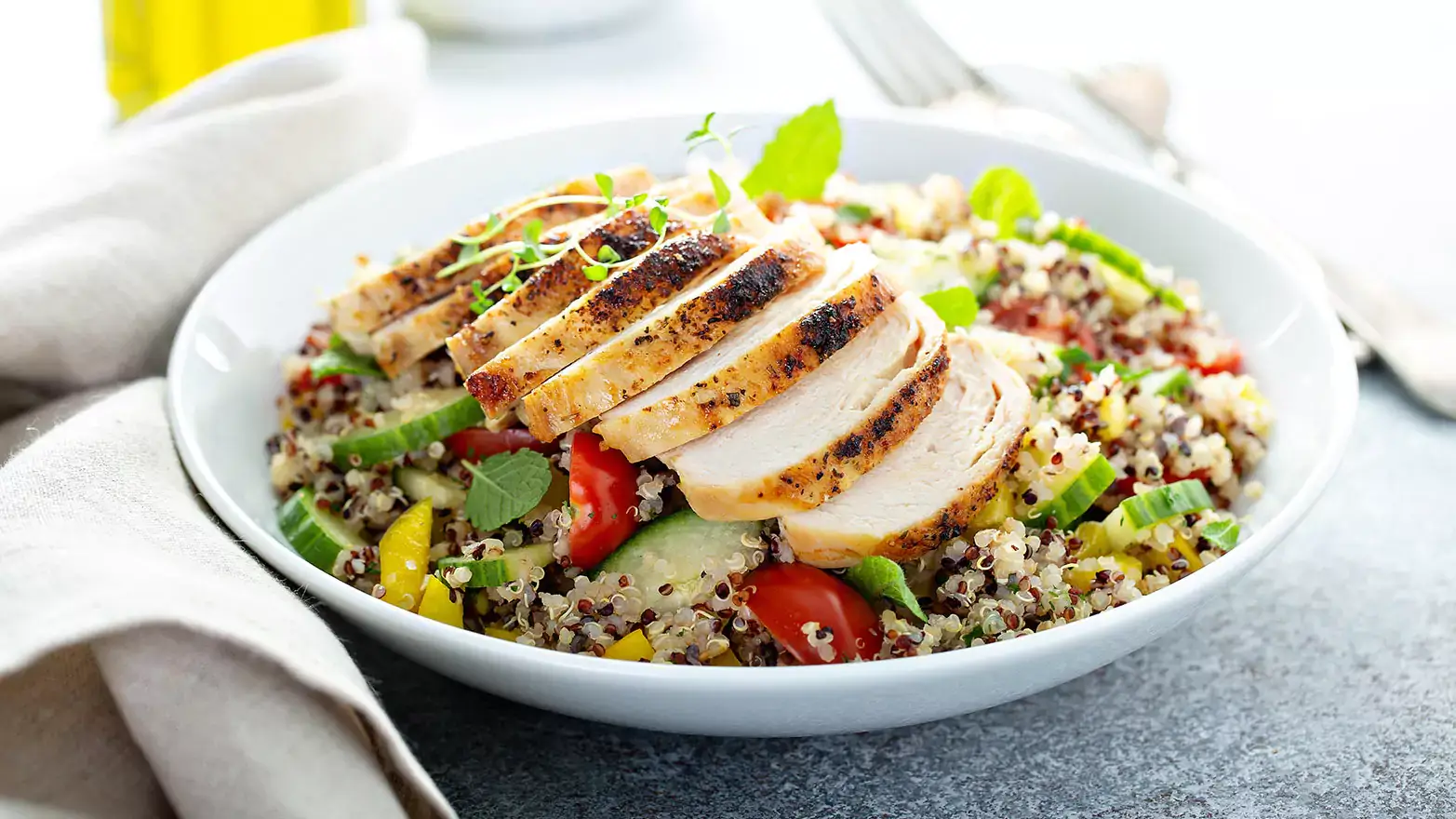 White bowl with quinoa, sliced cucumbers, cherry tomatoes, and sliced chicken breast next to a linen napkin