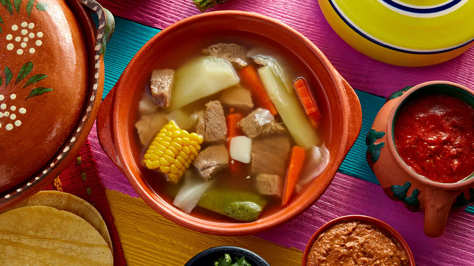 A bowl of Caldo de res on a colorful table spread.