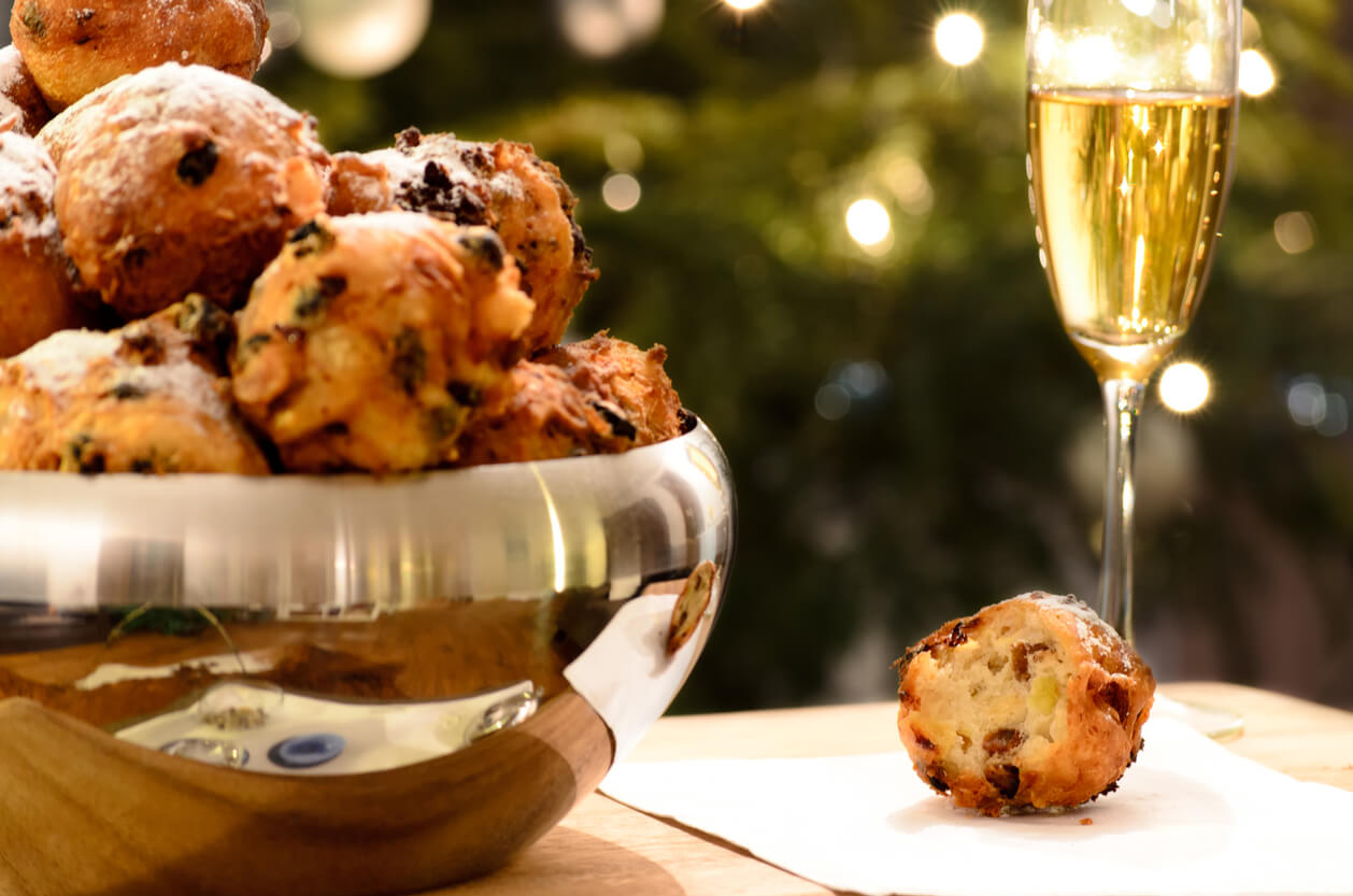 Oliebollen, oil balls or donut balls, a dutch pastry with raisins and powdered sugar traditionally eaten on New Year’s Eve in the Netherlands and a glass of champagne with a christmas tree in the background.