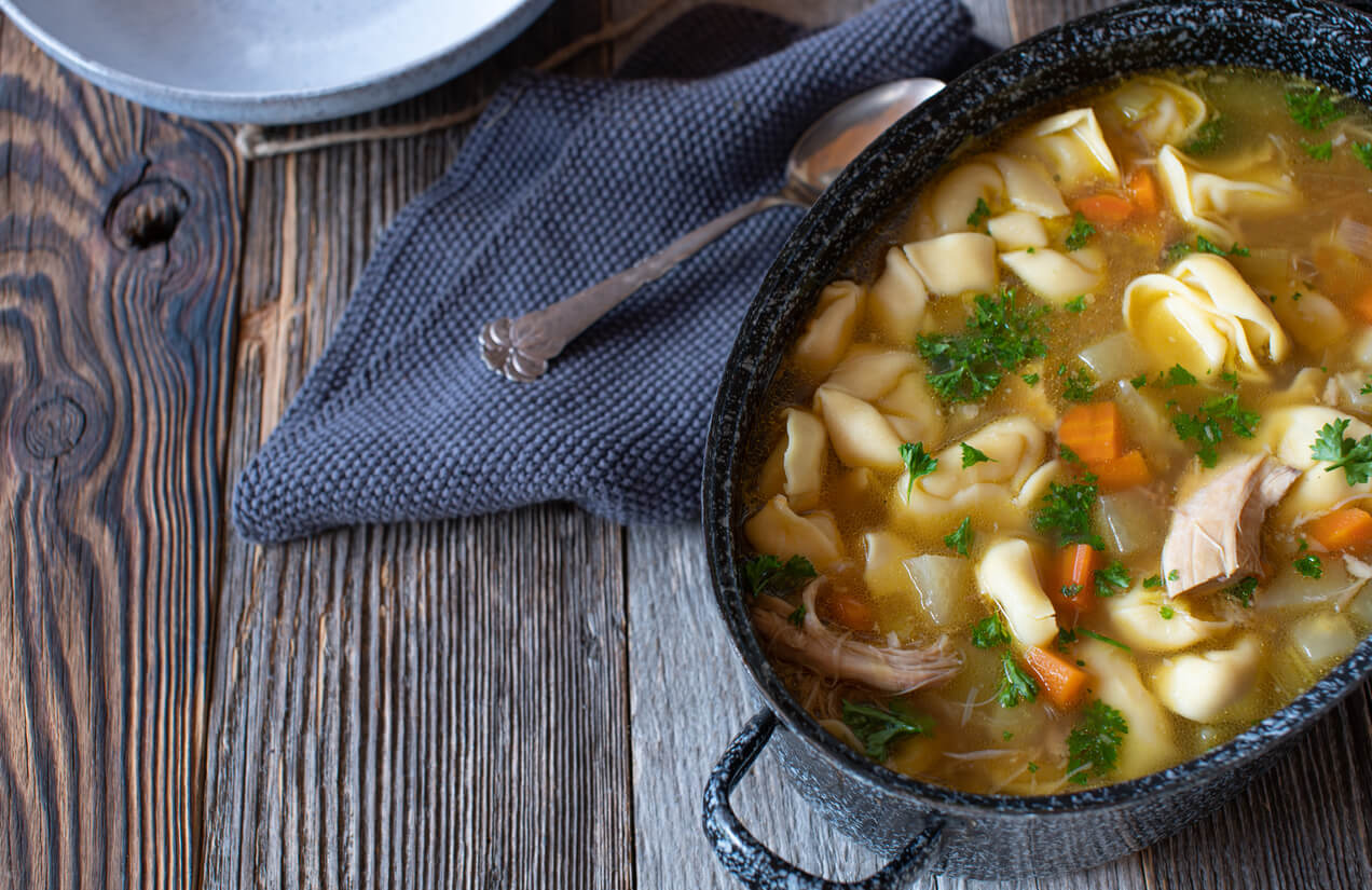 Homemade fresh cooked pot with chicken soup cooked with tortellini and root vegetable and served on wooden table background.