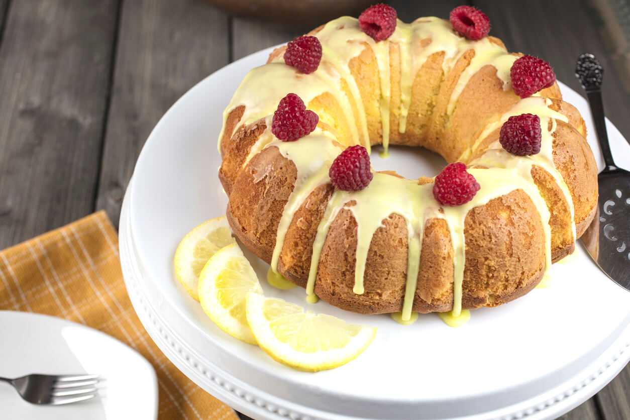 Lemon cake frosted with yellow sugar icing and red raspberries, dressed with lemon slices on the side on a white platter
