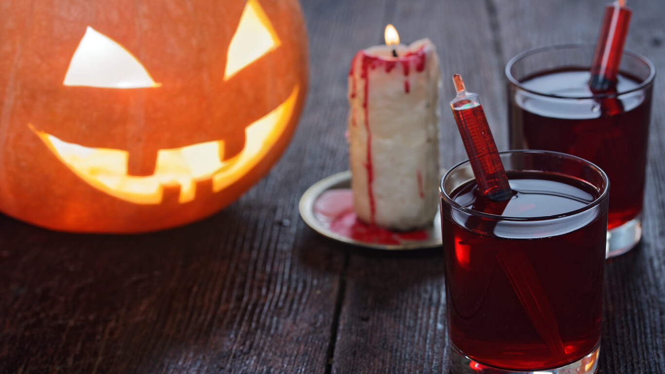 Halloween drink looking like blood served in glasses with syringes. Decoration with natural jack o'lantern and bloody candle. Close-up