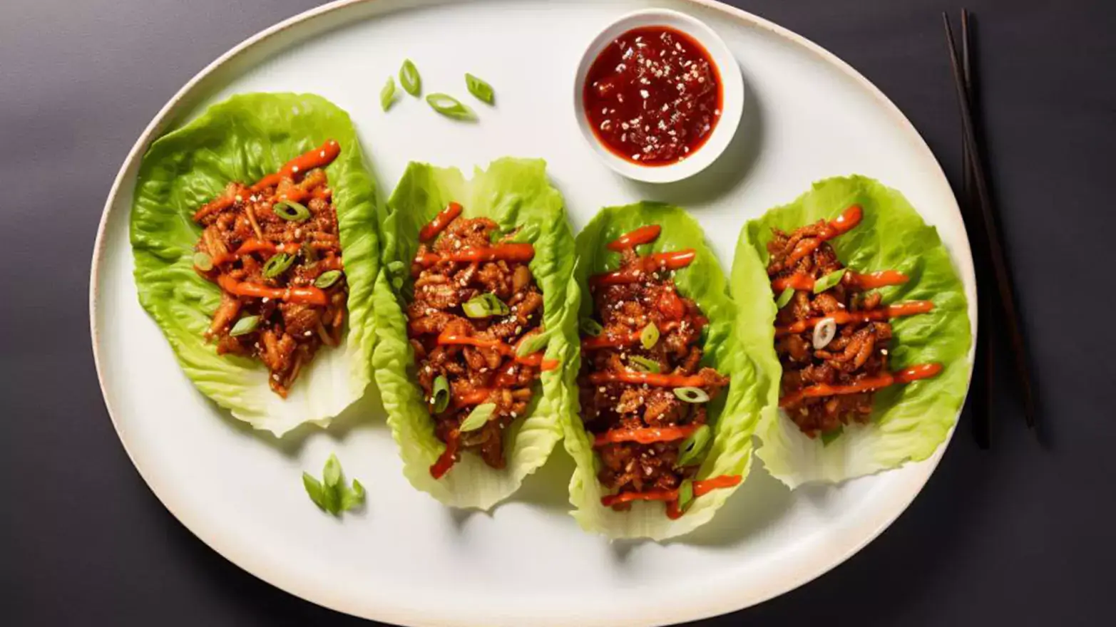 Large white plate on a black countertop with four lettuce cups holding stir fried chicken and scallions drizzled with sriracha next to a side of sriracha