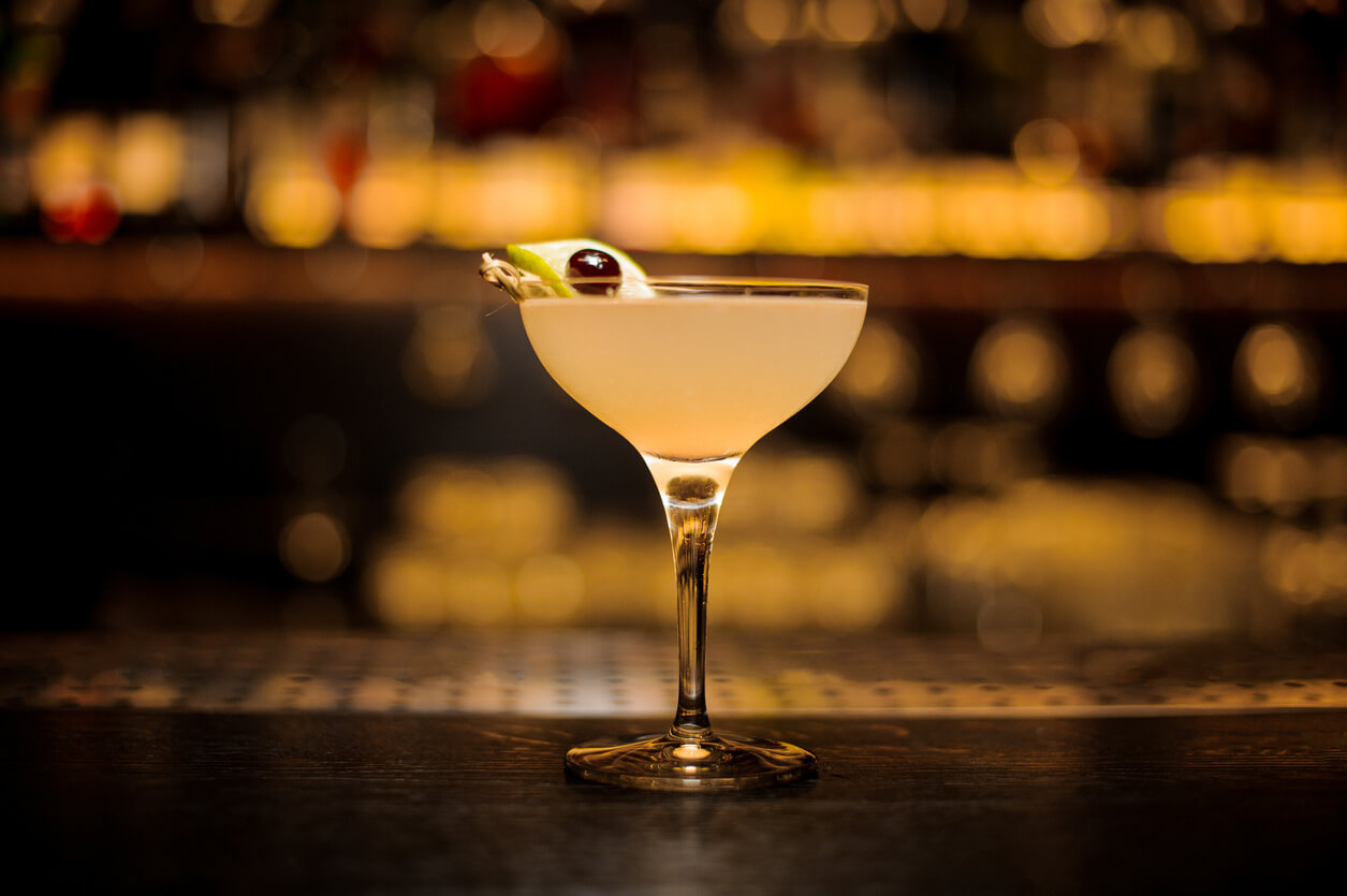 Glass of a Daiquiri cocktail decorated with lime slice and berry on a skewer standing on the bar counter on the blurred background.