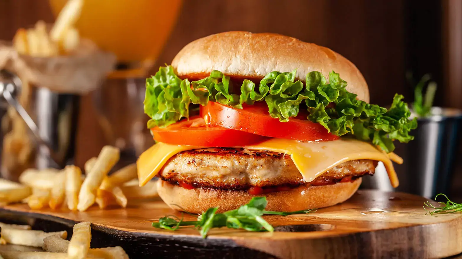 Two burger buns sandwiching a chicken patty, cheese, sliced tomatoes, and lettuce sitting on a wooden board next to french fries