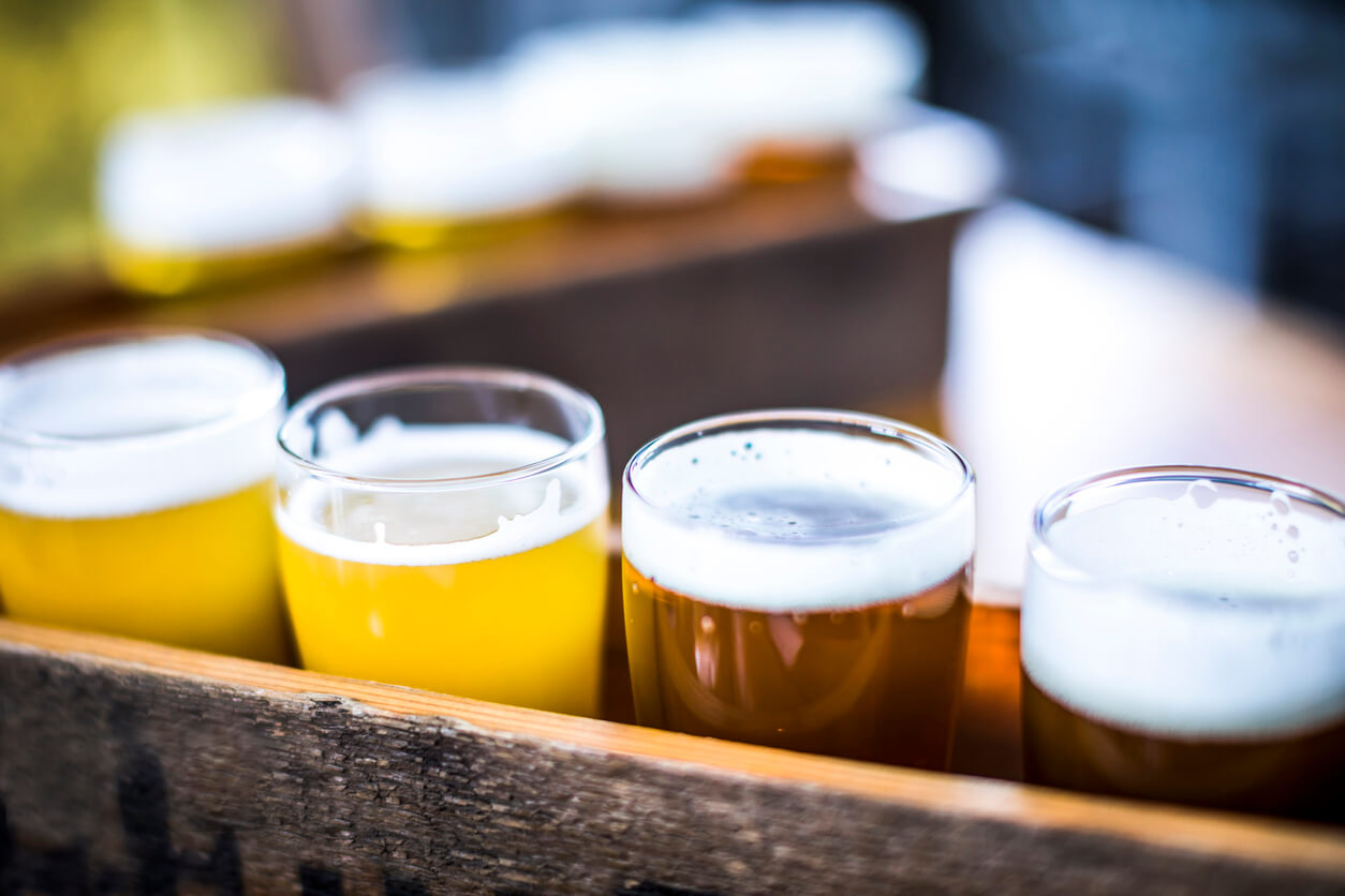 Flight of four beers lined up on a wooden table, with selective focus. Photo taken outdoors with natural lighting; beers range in color from light to very dark. Background out of focus.