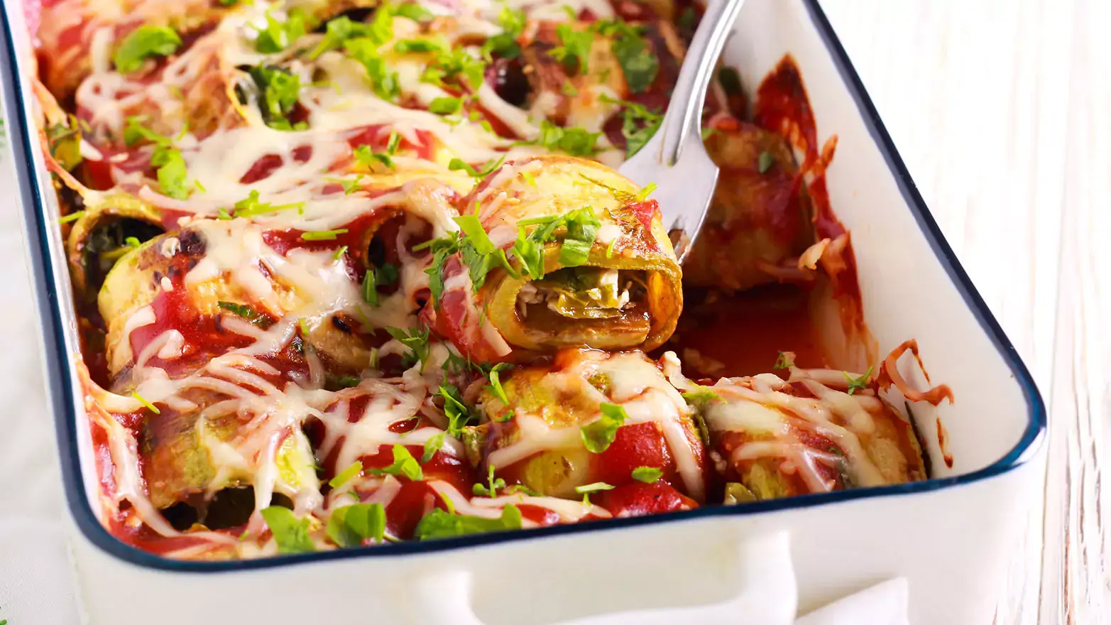 White casserole dish holding rolled-up zucchini strips topped with marinara sauce and shredded cheese