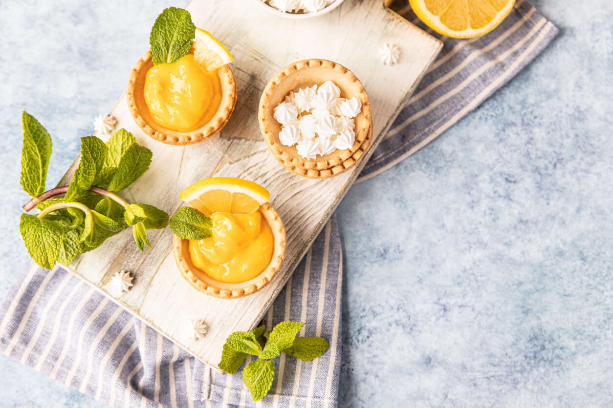 Shortbread tartlet filled with lemon curd, mint and lemon slices and mini meringue on wooden board, blue concrete background.