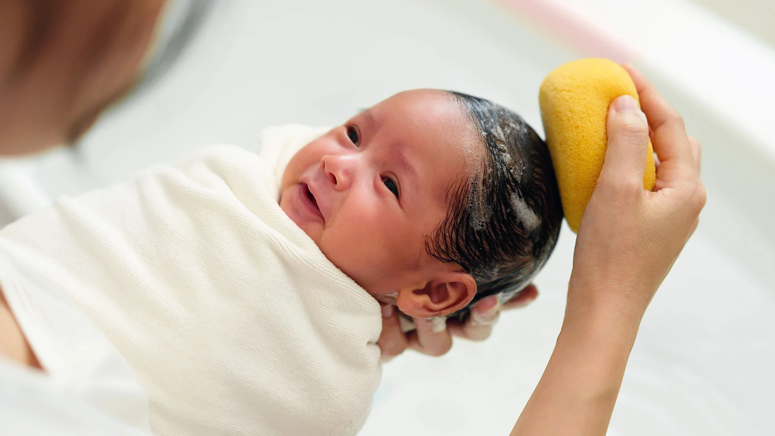 A stock photo of a baby in a bath, having their hair washed with a sponge.