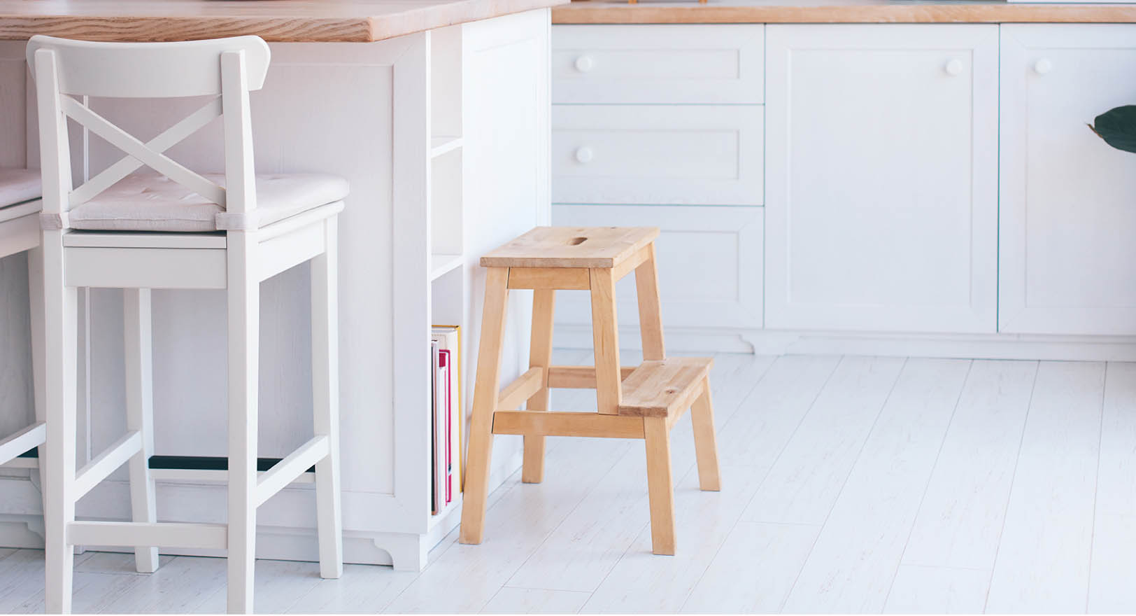 A photo of a wooden step stool on the floor next to a counter in a bright white kitchen.