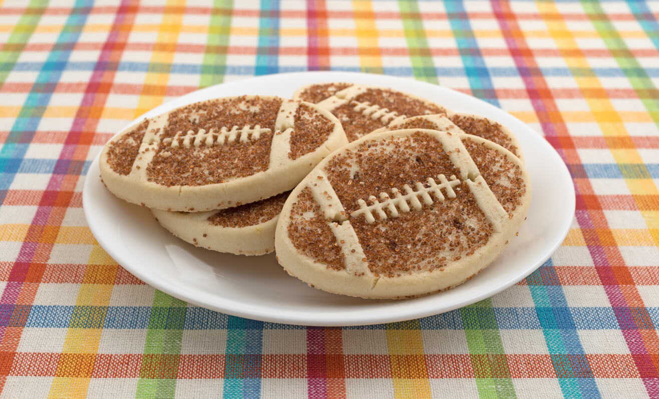 A plate full of football shaped sugar cookies with sprinkles on a colorful place mat.
