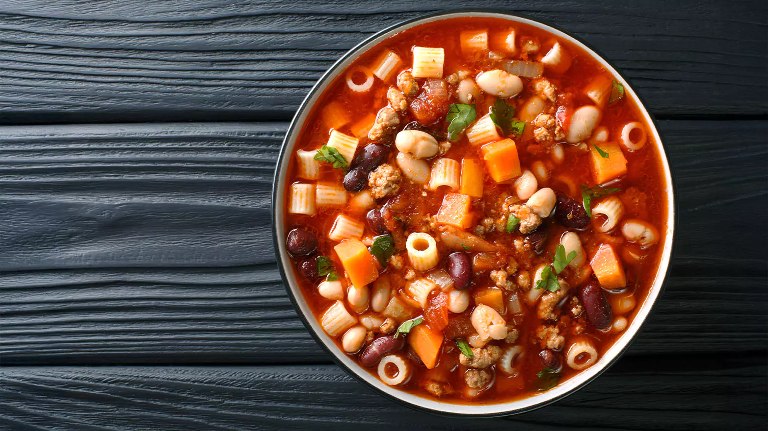 A bowl of ditalini soup with veggies and beans on a black wooden table.