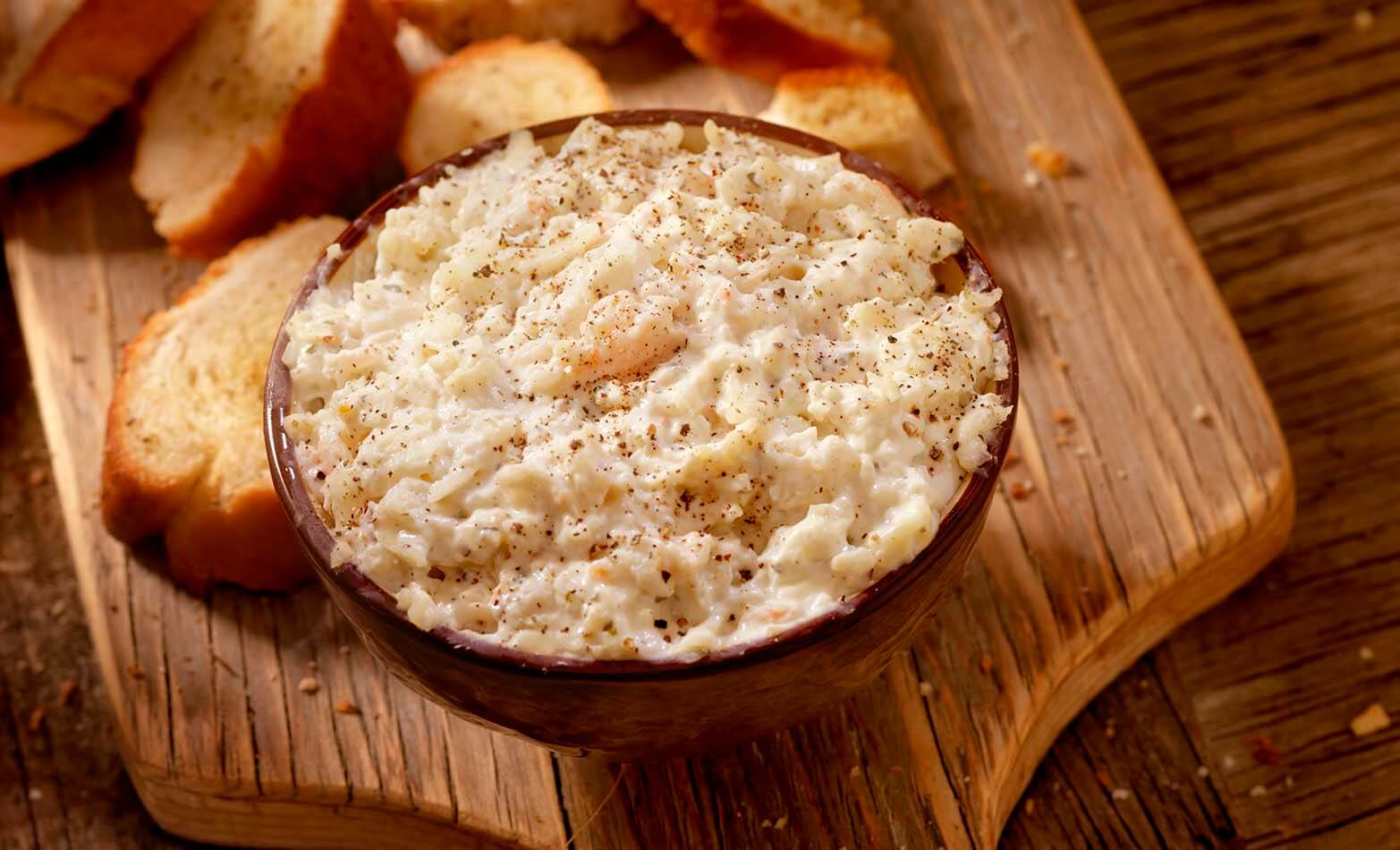 a bowl of creamy crab dip served with bread on a wooden cutting board