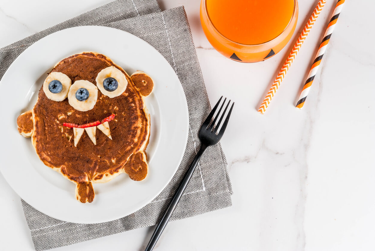 Funny food for Halloween. Kids breakfast pancake decorated like creepy monster, with banana, berries, with pumpkin smoothie juice.