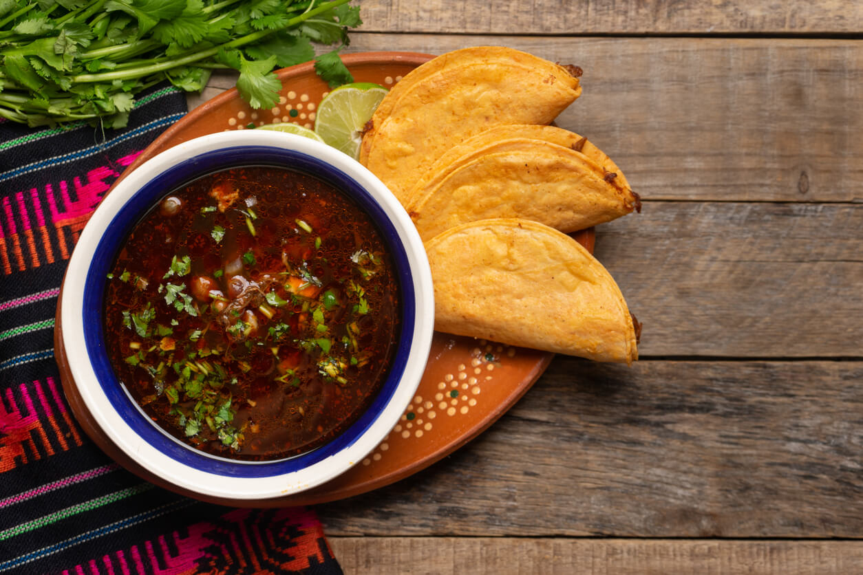 Traditional mexican birria consome soup and tacos on wooden background.