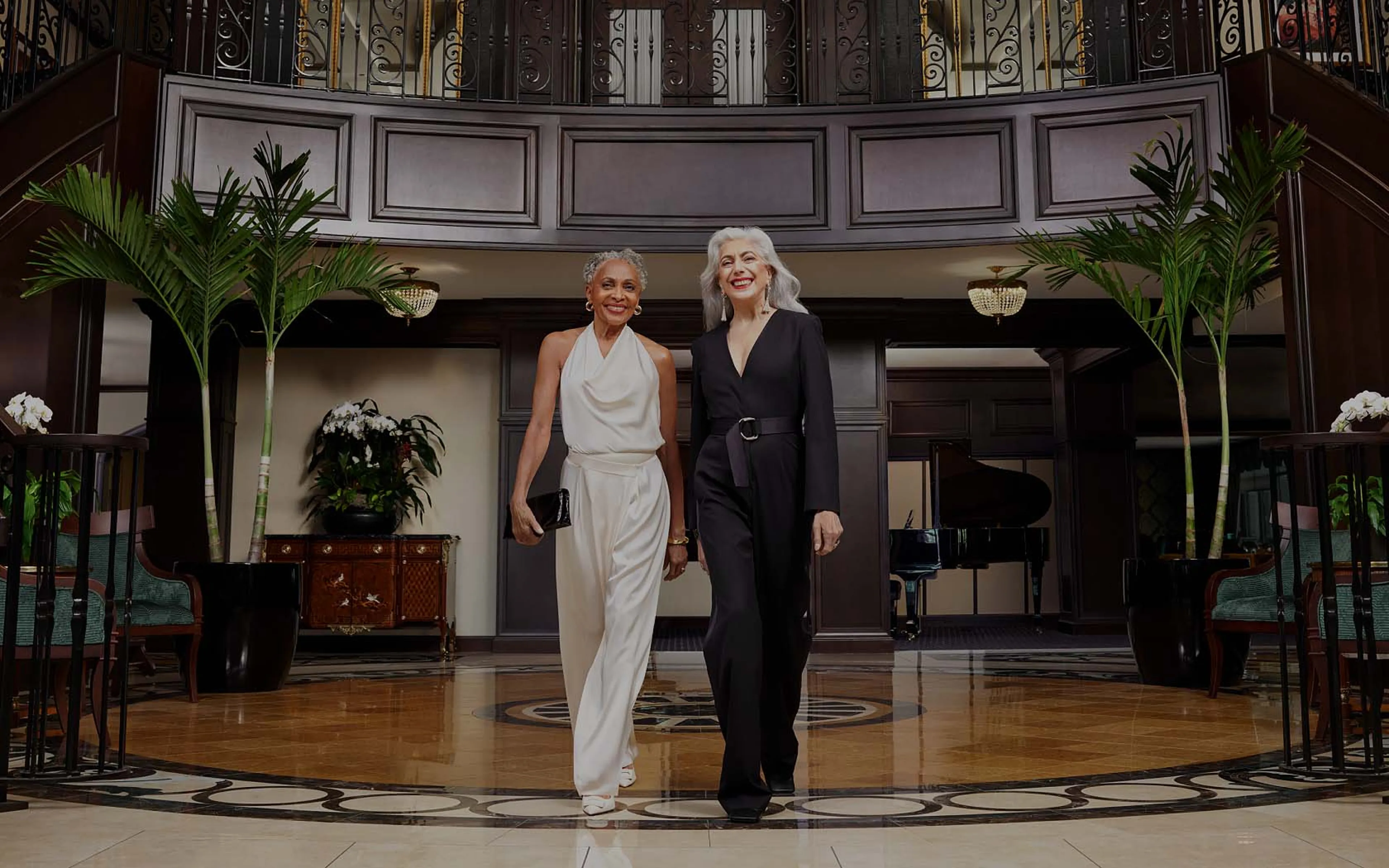 Two smiling women stand at the base of an elegant staircase in a luxurious lobby with a grand chandelier overhead.