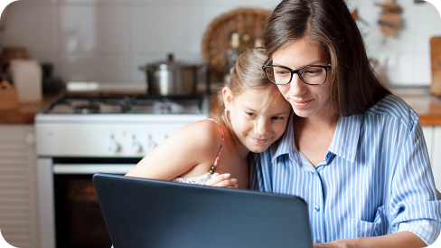 Mother wearing glasses and a blue button down shirt working on a laptop as her young daughter looks on