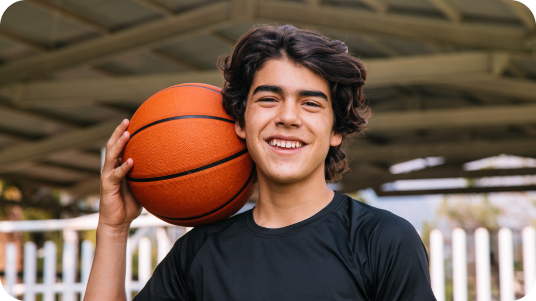 Teen boy with shaggy dark hair in a black t-shirt holding a basketball on his shoulder