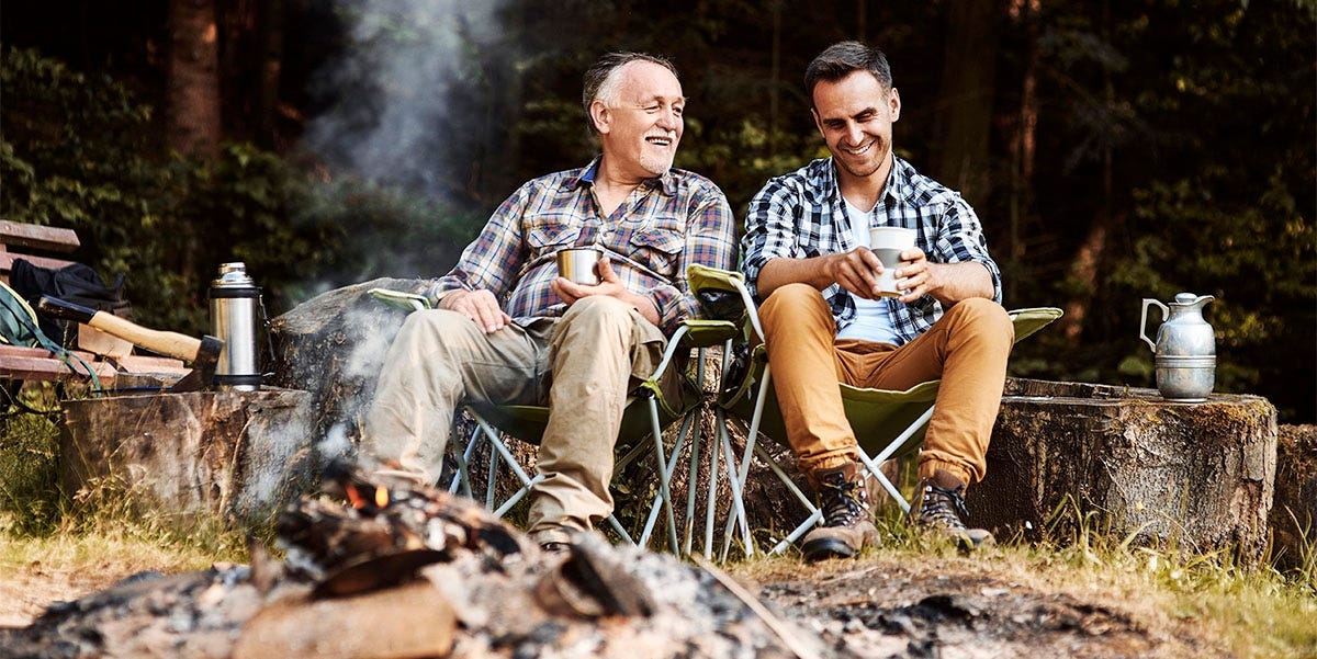 Elderly father sitting at a campfire with his son and drinking a hot bevarage together