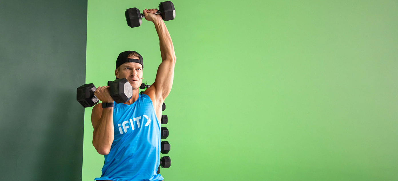 iFIT Trainer John Peel demonstrating proper form for a single-arm dumbbell overhead press in a bright fitness setting.