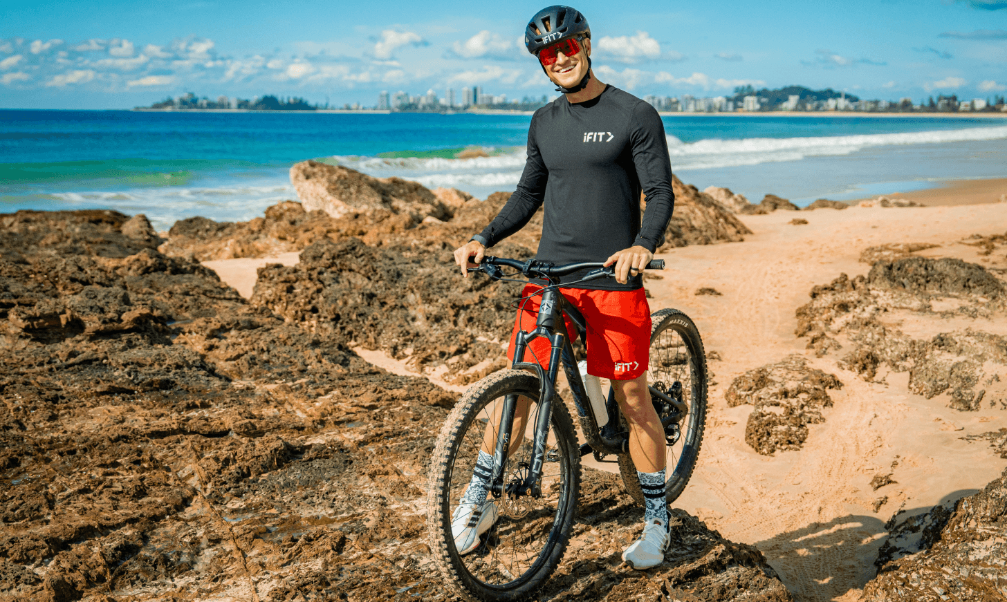iFIT Trainer John Peel stands with his bike on an Australian beach with a city-skyline in the distance.