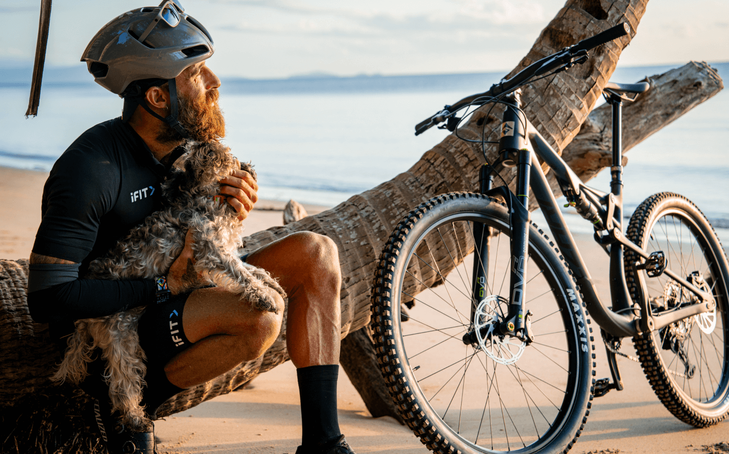 Tommy Rivs sits on a horizontal palm tree while holding a dog and looking out at the ocean.