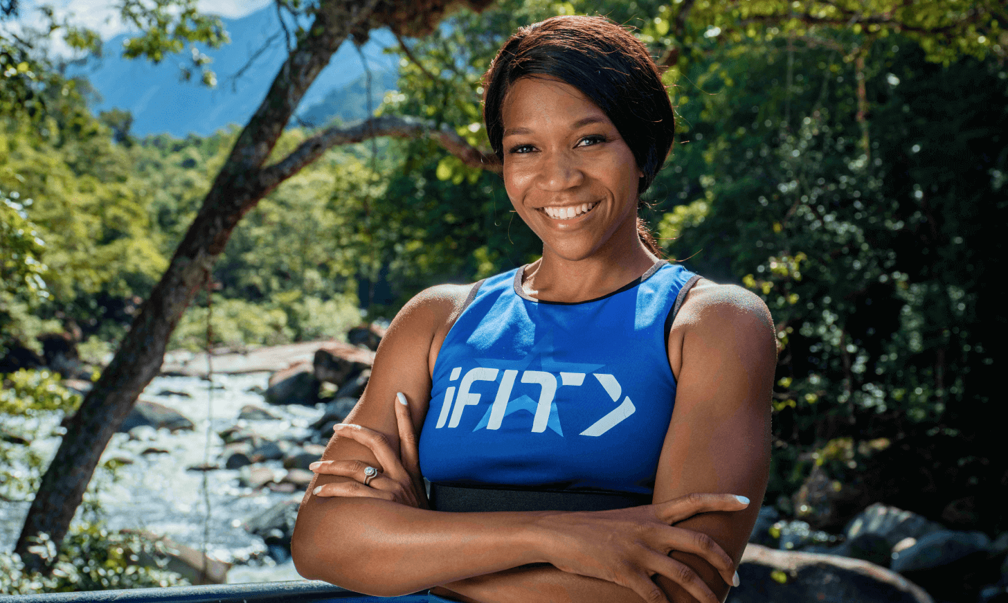 iFIT Trainer Shannon Cooper stands in front of nature-filled backdrop of trees, bushes, and a stream.