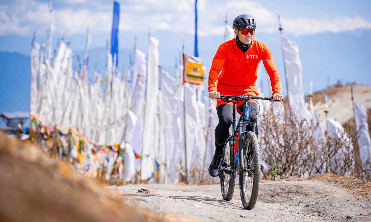 iFIT Trainer John Peel rides a bike past vertical white flags in Bhutan.