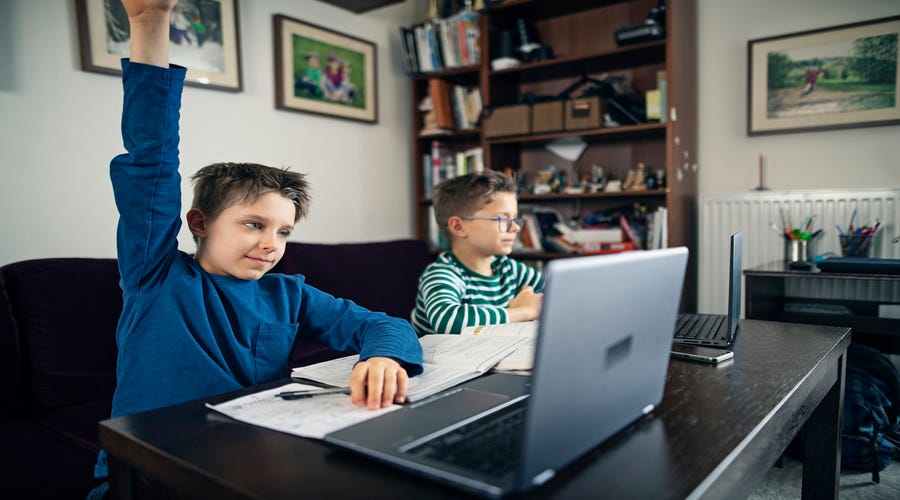 Zwei Jungen sitzen mit Laptop an einem Tisch, einer von ihnen hebt die Hand und zeigt auf.