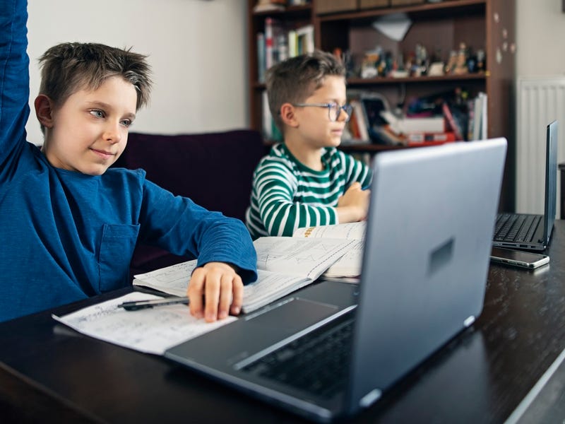 Zwei Jungen sitzen mit Laptop an einem Tisch, einer von ihnen hebt die Hand und zeigt auf.
