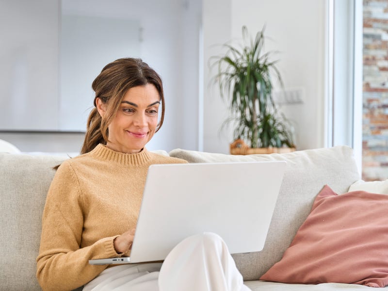 Frau sitzt auf einem Sofa mit ihrem aufgeklappten Laptop auf dem Schoß.