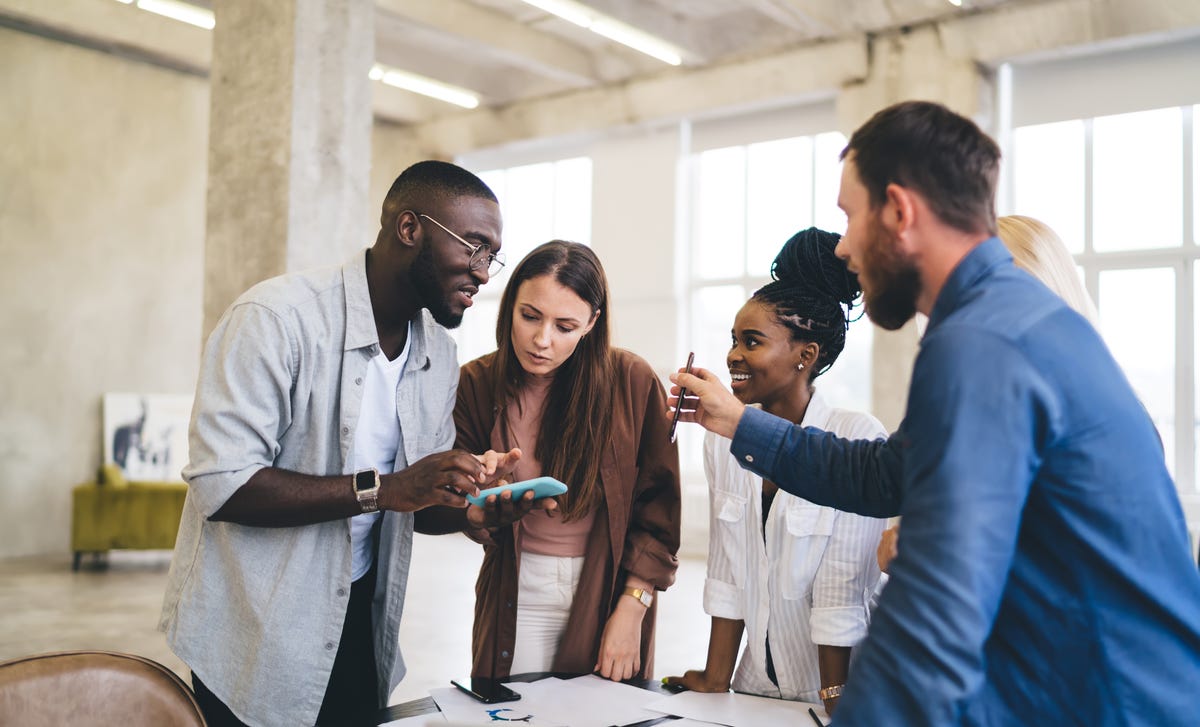 Eine Gruppe von 4 Leuten unterschiedlicher Nationen spricht miteinander. Zwei Männer, zwei Frauen. Ein Mann zeigt was an seinem Handy.